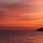 Sunset over calm ocean with an orange-pink sky and a dark hillside on the right.