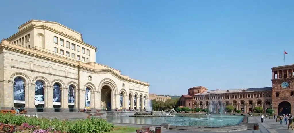 Large stone building with arches and a central entrance beside a circular fountain in a sunlit plaza with pedestrians nearby.
