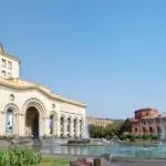 Large stone building with arches and a central entrance beside a circular fountain in a sunlit plaza with pedestrians nearby.