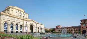 Large stone building with arches and a central entrance beside a circular fountain in a sunlit plaza with pedestrians nearby.