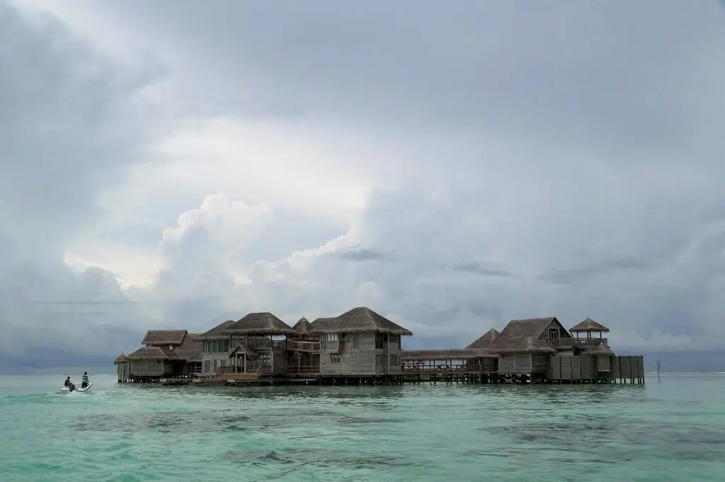 Overwater bungalows with thatched roofs sit on calm turquoise water under a cloudy sky.