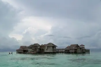 Overwater bungalows with thatched roofs sit on calm turquoise water under a cloudy sky.
