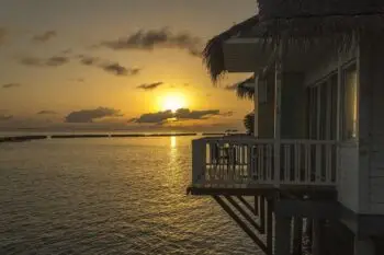 Sunset over the calm sea with a thatched-roof bungalow on stilts and a wooden balcony above the water.