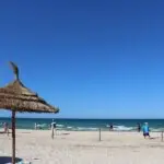 Sunny beach scene with straw umbrellas, people on the sand and calm waves under a clear blue sky.