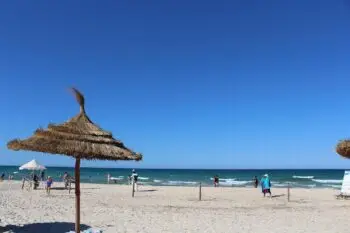 Sunny beach scene with straw umbrellas, people on the sand and calm waves under a clear blue sky.