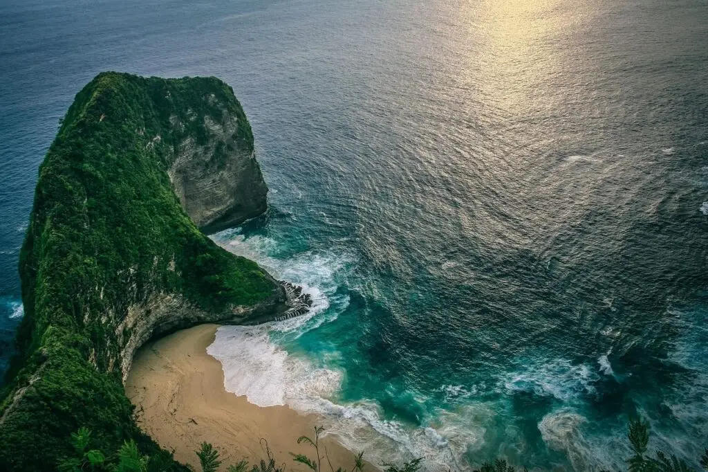 Aerial view of a green, cliff-like rock forming a cove with a sandy beach and turquoise water
