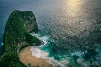 Aerial view of a green, cliff-like rock forming a cove with a sandy beach and turquoise water