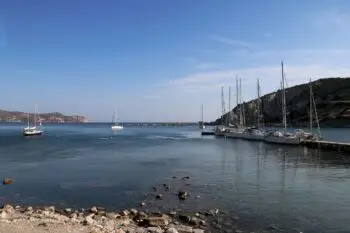 Calm coastal harbor with several sailboats docked at a rocky pier under a clear blue sky.