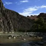 Rugged river canyon with steep rocky cliffs on the left, calm water and scattered rocks along the shore, and green trees on the right under a blue sky