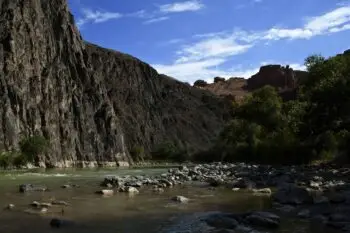 Rugged river canyon with steep rocky cliffs on the left, calm water and scattered rocks along the shore, and green trees on the right under a blue sky