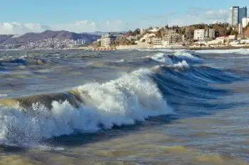 Waves crash near a sandy shore with a city skyline in the distance on a clear day