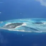 Aerial view of a tropical island resort with white sand beaches, palm trees, and overwater bungalows in a turquoise lagoon.