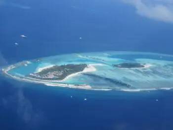 Aerial view of a tropical island resort with white sand beaches, palm trees, and overwater bungalows in a turquoise lagoon.