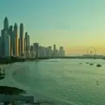 Wide city skyline along a calm bay at sunset, with a sandy beach and boats on the water.