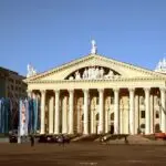 Neoclassical theater with tall columns, statues on the pediment, and colorful flags along the plaza under a clear blue sky.