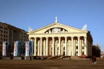 Neoclassical theater with tall columns, statues on the pediment, and colorful flags along the plaza under a clear blue sky.