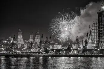 Nighttime harbor scene with fireworks over a city skyline and construction cranes along the water at night (B&W).