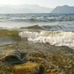 Clear shallow water over a pebbled shore with small waves and distant hazy mountains on the horizon.