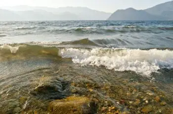 Clear shallow water over a pebbled shore with small waves and distant hazy mountains on the horizon.