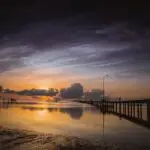 Sunset over a calm sea with a wooden pier extending into the water; a silhouetted figure stands at the far end.
