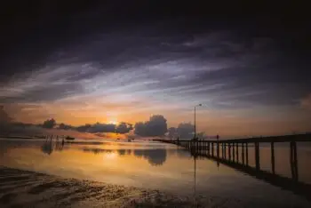 Sunset over a calm sea with a wooden pier extending into the water; a silhouetted figure stands at the far end.