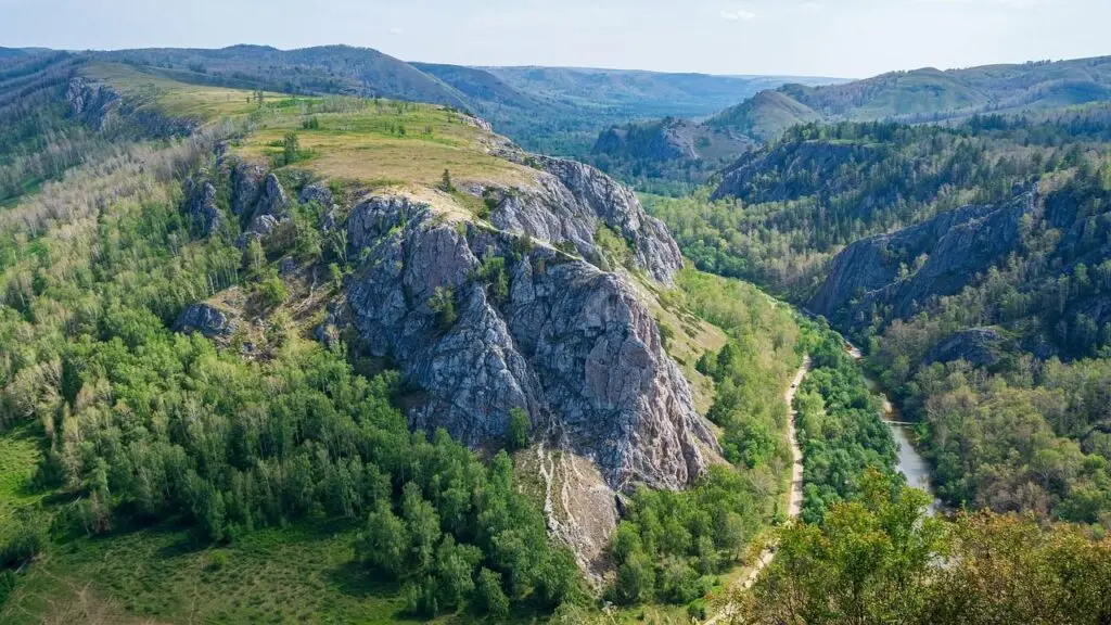 Aerial view of a rocky hill surrounded by green trees, with a narrow dirt path winding along the ridge and a river in the valley nearby