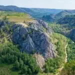 Aerial view of a rocky hill surrounded by green trees, with a narrow dirt path winding along the ridge and a river in the valley nearby