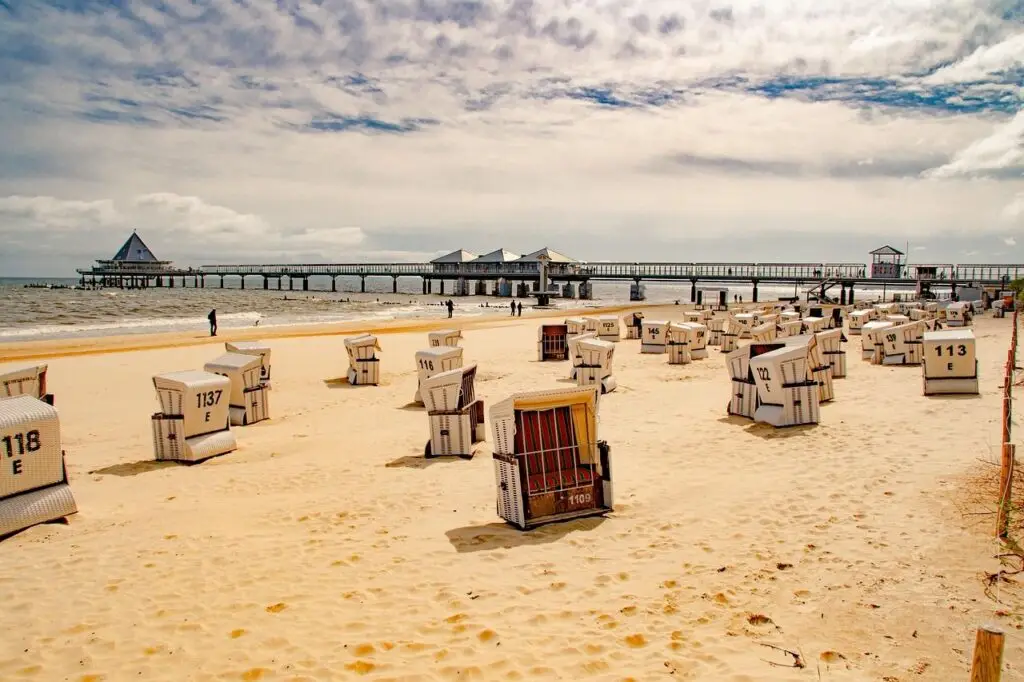 Sunlit beach with many white striped beach chairs facing the sea and a wooden pier extending into the water.