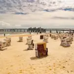 Sunlit beach with many white striped beach chairs facing the sea and a wooden pier extending into the water.