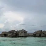Overwater thatched-roof bungalows on stilts above a turquoise lagoon under a cloudy sky.