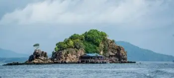Small rocky island with lush trees and wooden huts built on stilts over calm blue water.