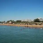 Busy beach with swimmers in calm blue water, sandy shore lined with straw umbrellas and palm trees, and white resort buildings in the background.