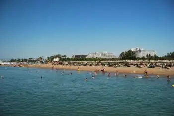 Busy beach with people swimming and sunbathing, rows of thatched umbrellas and white hotel buildings in the background.