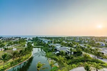 A coastal resort with modern white villas, palm trees, and a curved canal reflecting the sky at sunset, overlooking the ocean.