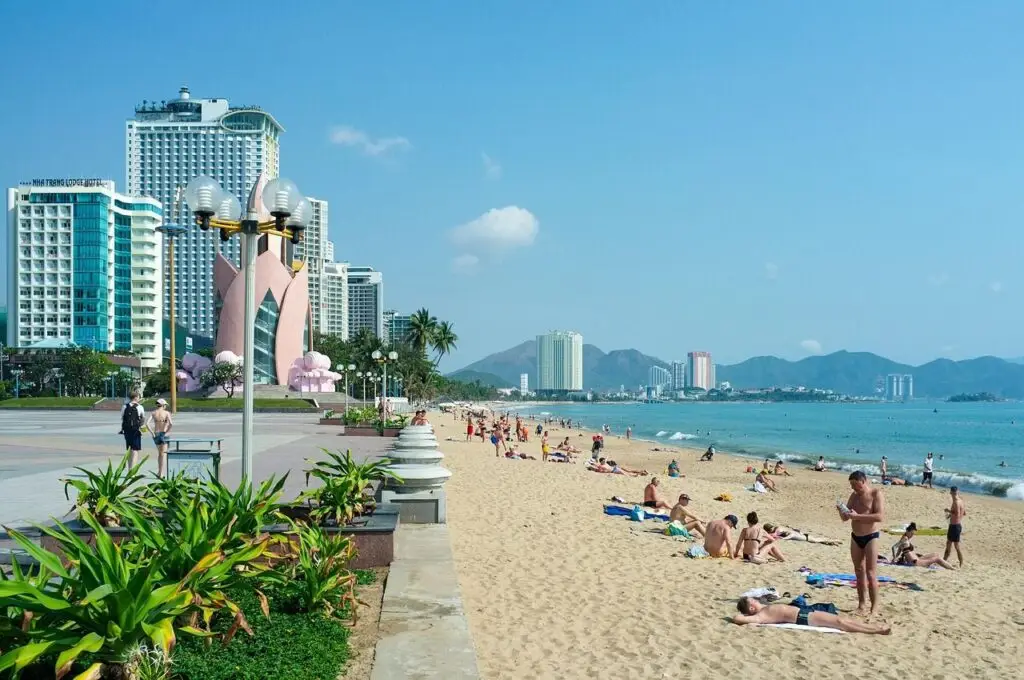 Sunny beach with people sunbathing along a promenade and modern high-rise buildings lining the shore.
