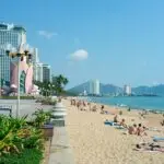 Sunny beach with people sunbathing along a promenade and modern high-rise buildings lining the shore.