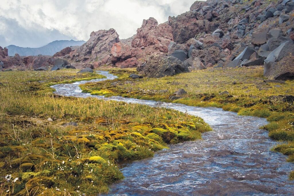 A narrow stream winds through mossy grassland with jagged rocky hills in the background under a cloudy sky.