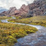 A narrow stream winds through mossy grassland with jagged rocky hills in the background under a cloudy sky.