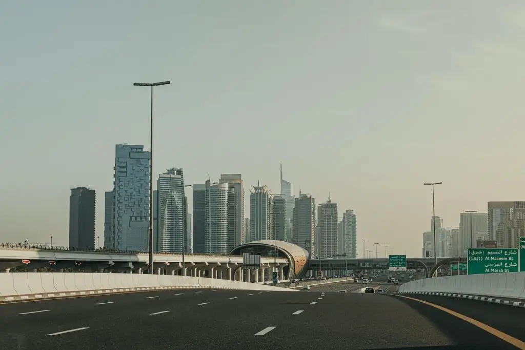 Wide highway with white barriers leads toward a cluster of modern skyscrapers under a clear sky.",