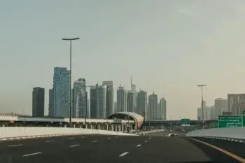 Wide highway with white barriers leads toward a cluster of modern skyscrapers under a clear sky.",